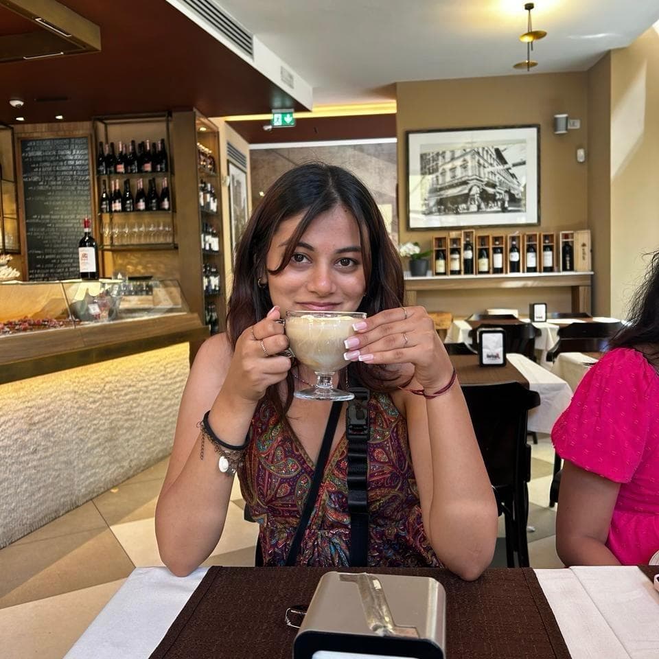 Bhavi holding a cup of coffee at a café table, smiling at the camera.