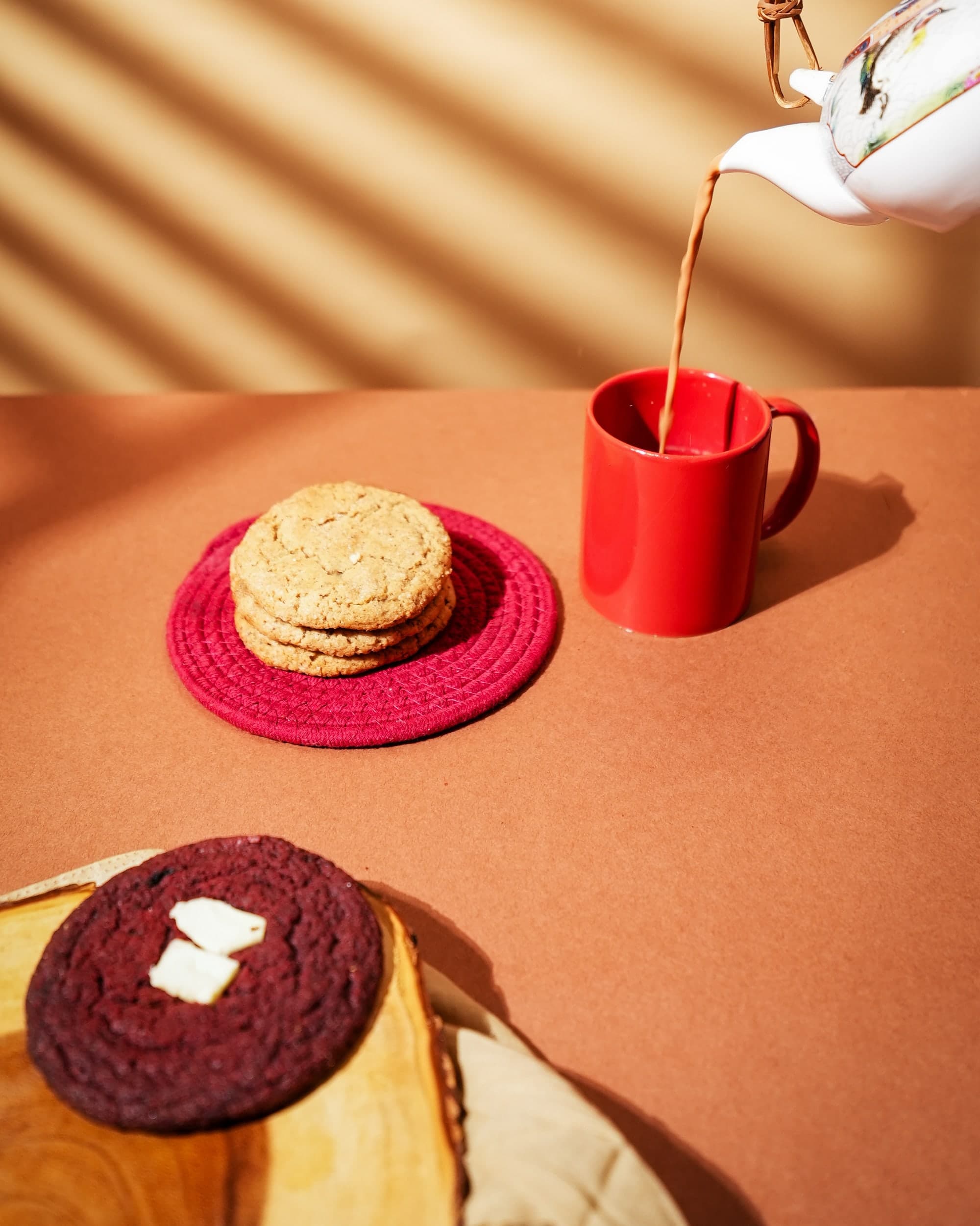 A stack of Chai Latte cookies on a red mat, beside chai being poured into a red mug from a floral teapot.