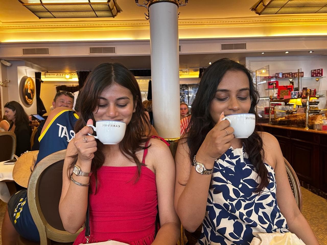 Bhavi and Kriti drinking coffee side by side at a café, cups raised.