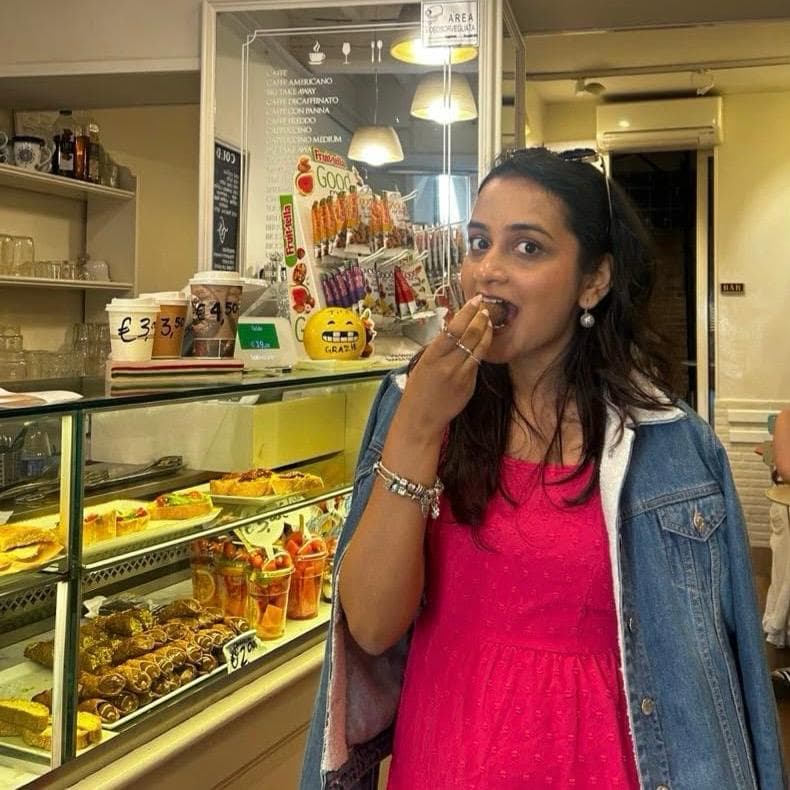 Kriti taking a bite of a pastry at a café counter, a lit glass case of cannoli and pastries beside her.