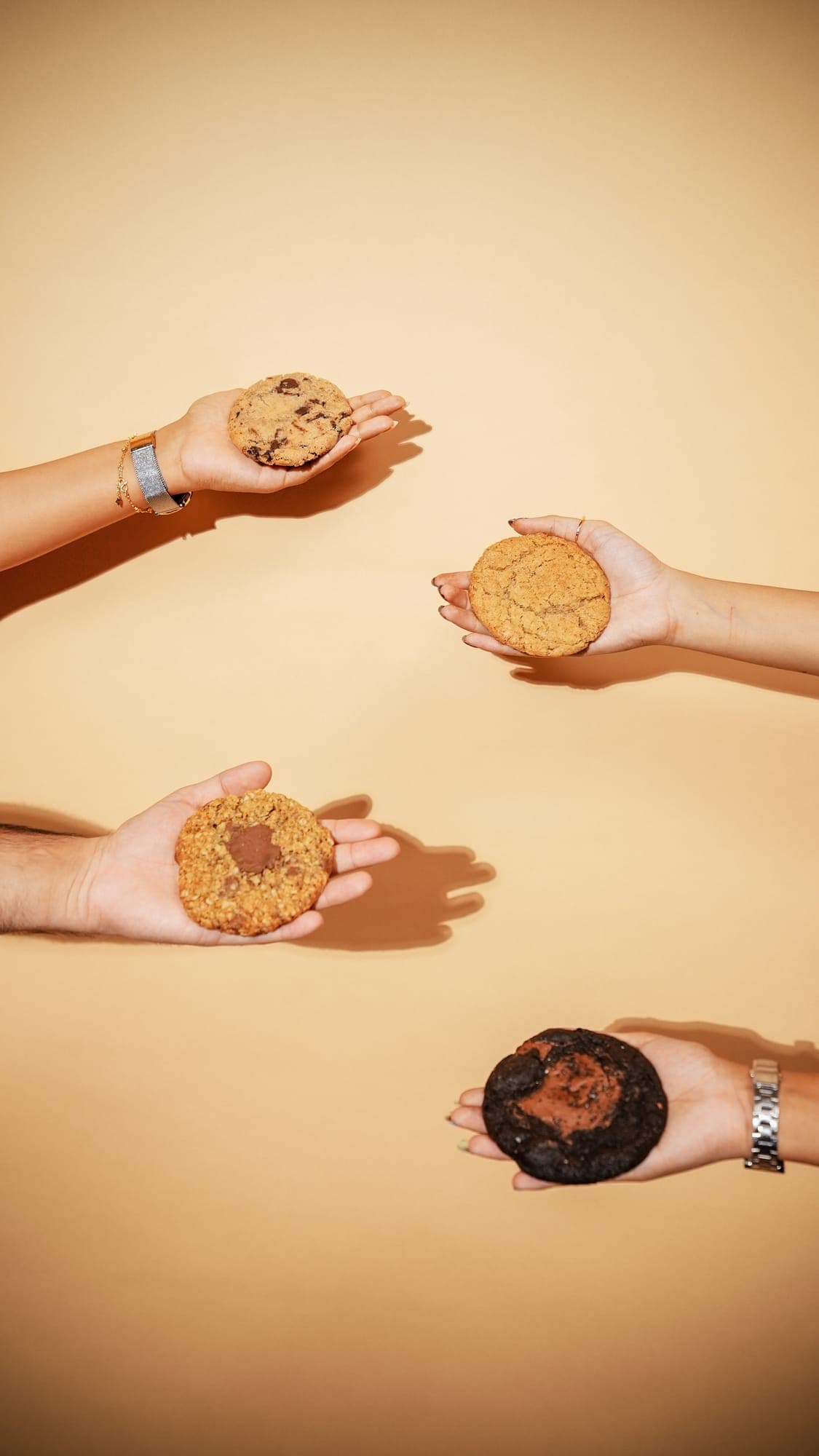 Four hands, each holding out a different cookie against a warm tan background.